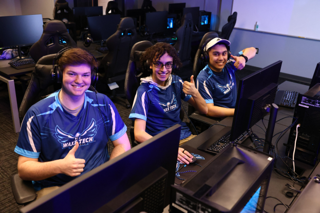 Drei junge Männer mit Headsets lächeln, während sie an einem Schreibtisch mit Computern, Monitoren, Tastaturen und Kabeln sitzen, was darauf hindeutet, dass sie ein Spiel als Teil eines E-Sport-Teams spielen.