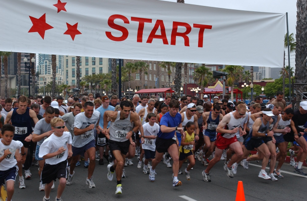 Eine Gruppe von Menschen, die bei einem Marathon laufen, mit einem Verkehrskegel im Vordergrund und einem Banner mit Text im Hintergrund, vor Bäumen, Laternenpfählen, Gebäuden und einem klaren blauen Himmel.