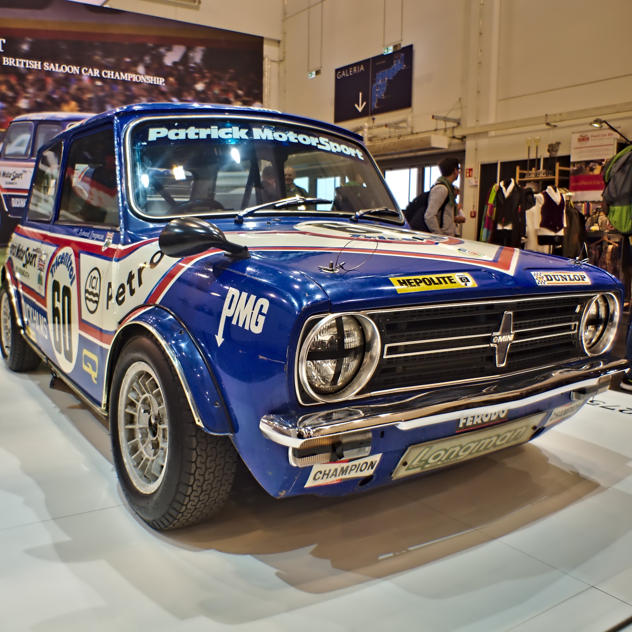 Ein kleiner blauer und weißer Mini Cooper S Rallye-Wagen in einem Museum ausgestellt, umgeben von Menschen mit Taschen.