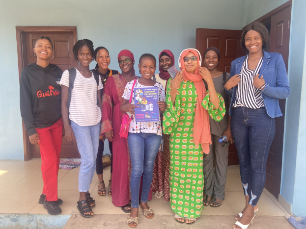 Eine Gruppe von Frauen, die vor einem Schulgebäude in Nigeria Bücher halten und lächeln, mit einem Tuch auf dem Boden und sichtbaren Türen im Hintergrund.