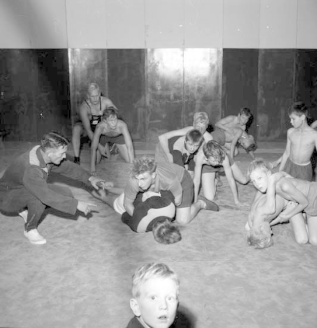 Schwarz-weißes Bild von jungen Jungs, die in einem Fitnessstudio miteinander ringen, mit einer Wand im Hintergrund.