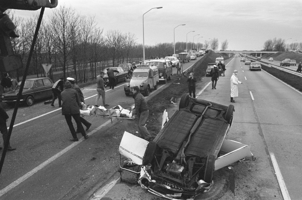 Schwarz-weißes Bild eines umgestürzten Autos am Straßenrand mit beschädigten Fahrzeugen in der Nähe und einer Gruppe von Menschen drumherum, vor dem Hintergrund von Laternen, Bäumen, einer Brücke und dem Himmel.