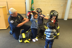 Eine Gruppe von Kindern in Feuerwehranzügen und ein Mann in einem Feuerwehranzug stehen neben einer Feuerwache im Inneren, wobei der Mann einen Helm und eine Tasche auf dem Boden hält.