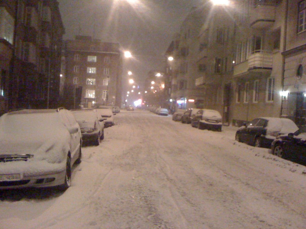Schneebedeckte Kraftfahrzeuge auf einer Straße mit Laternen, Straßenlaternen, Gebäuden und einem Himmel im Hintergrund.