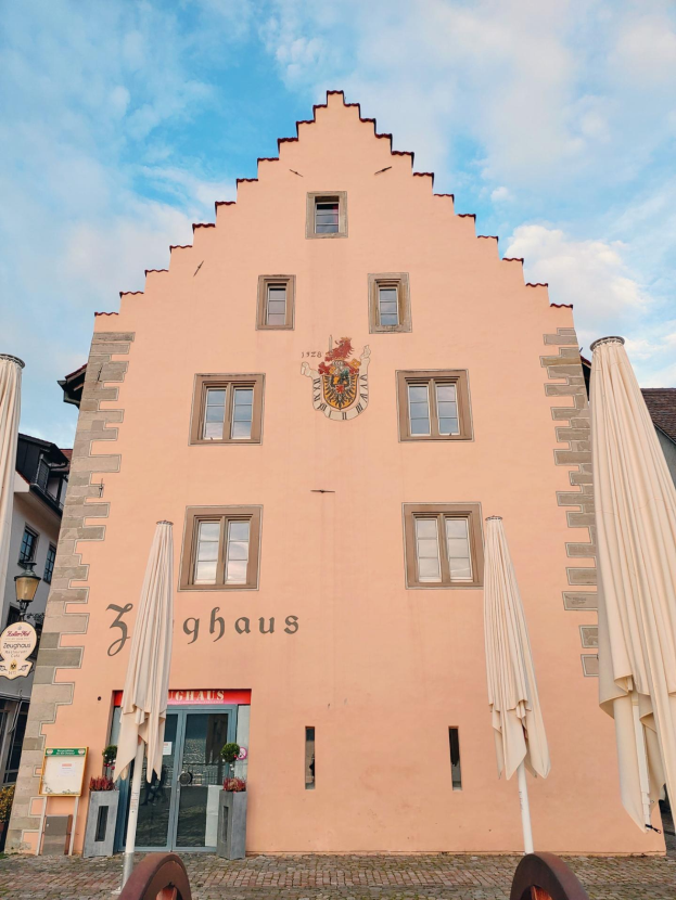 Ein rosafarbenes Gebäude mit einem zentralen Uhrenturm, umgeben von Fenstern, Schirmen, einem Schild, einer Straßenlaterne, Topfpflanzen, unter einem bewölkten Himmel, repräsentiert München, Deutschland.