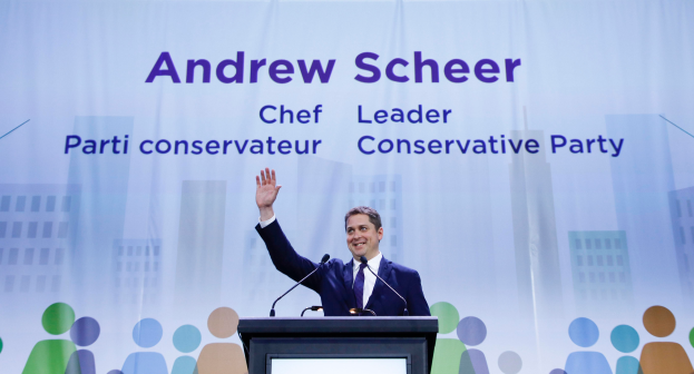 Andrew Scheer, der Vorsitzende der Conservative Party, steht vor einem Podium mit zwei Mikrofonen und einem Bildschirm, mit einem Banner im Hintergrund, das Text und Gebäudeabbildungen zeigt.