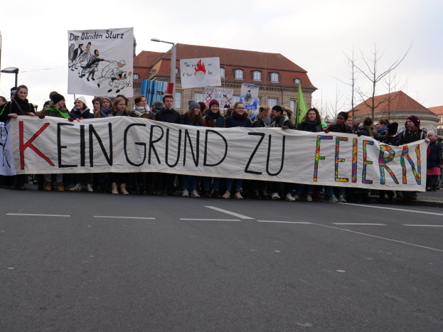 Demonstranten halten ein Transparent mit der Aufschrift "Kein Grund zu Feiern" gegen Sparmaßnahmen, mit Straßeninfrastruktur und Gebäuden im Hintergrund unter einem klaren Himmel.
