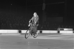 Schwarz-weiß-Foto eines Mannes, der auf einem Eisstadion Fahrrad fährt, mit Zuschauern im Hintergrund und einer Wand dahinter.