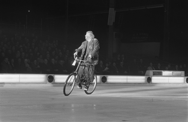 Schwarz-weiß-Foto eines Mannes, der auf einem Eisstadion Fahrrad fährt, mit Zuschauern im Hintergrund und einer Wand dahinter.
