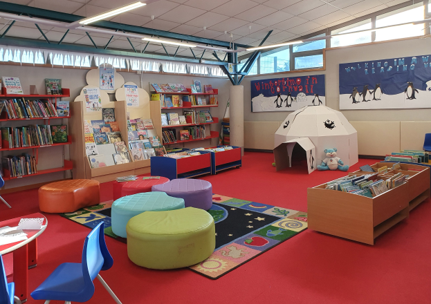 Eine hell erleuchtete Kinderbibliothek mit Bücherregalen, Tischen, Stühlen, Teppichen, Wandtafeln und Glasfenstern.