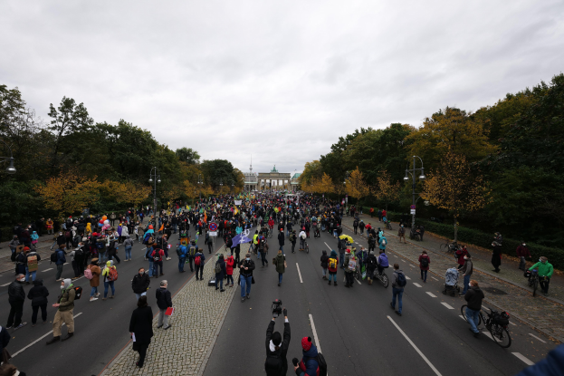 Eine große Gruppe von Menschen marschiert eine von Bäumen gesäumte Straße in Berlin entlang und hält Kameras, mit einem Gebäude und einem klaren Himmel im Hintergrund.