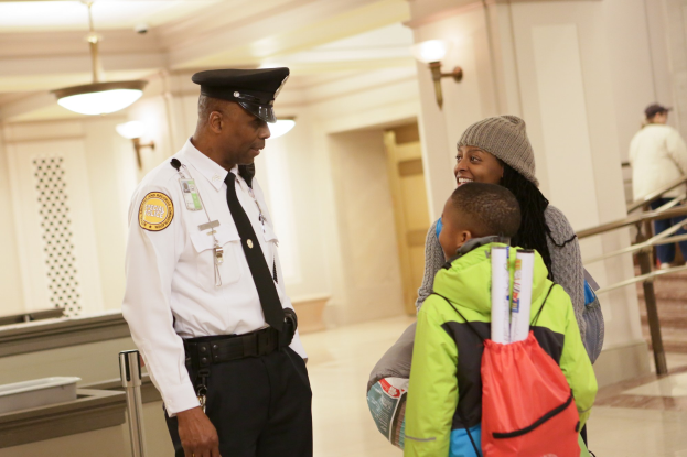 Ein Polizeibeamter spricht mit einer Frau in einer Mütze und einem Kind an einem Flughafen, mit anderen Personen im Hintergrund neben Treppen und wandmontierten Lampen.