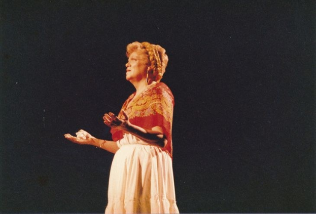 Eine Frau in einem weißen, hochgeschlossenen, langärmligen Kleid steht auf einer dunklen Bühne mit ausgestreckten Händen, ihr Haar ist zu einem Dutt gebunden und ihr Gesichtsausdruck ernst.