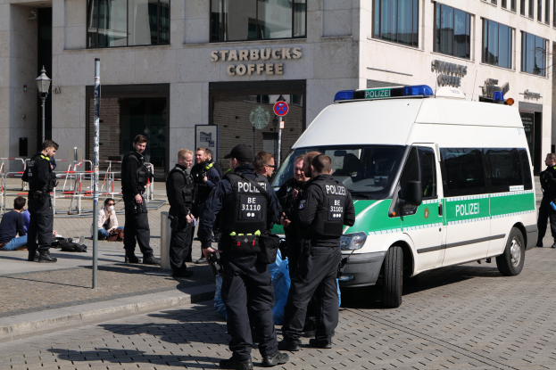 Polizeibeamte vor einem Starbucks-Café mit einem Van auf der rechten Seite und Menschen auf der linken Seite, vor einem Hintergrund aus Gebäude, Schilder, Laterne und Zaun.