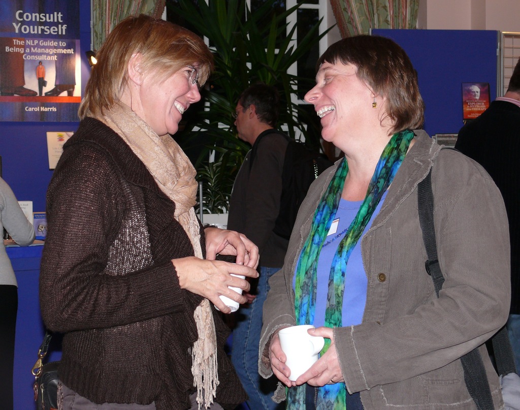 Zwei Frauen stehen sich gegenüber und lachen, jede hält einen weißen Becher, mit einem Mann, der neben einer Pflanze am Fenster steht, und zwei weiteren Personen und Plakaten im Hintergrund.