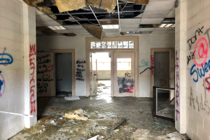 Innenansicht eines verlassenen Gebäudes mit graffitibedeckten Wänden und Decke, Schutt auf dem Boden, mehreren Türen und Fenstern und verstreuten Gegenständen.