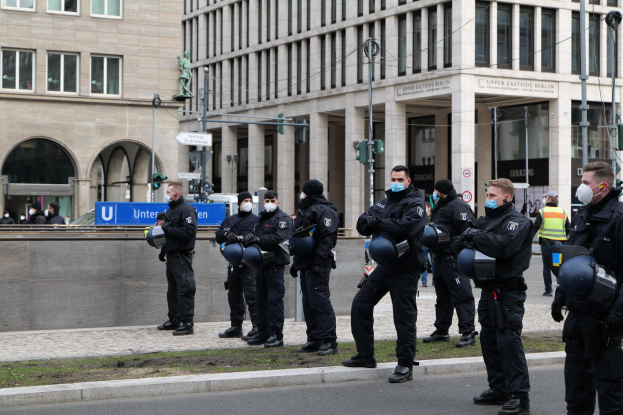 Polizeibeamte in schwarzen Uniformen und Masken vor einem Berliner Gebäude mit Glasfenstern und Säulen, einige halten Helme, mit Laternenmasten, Verkehrsampeln, Schildern mit Text, einer Statue und anderen Menschen im Hintergrund.