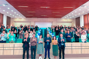 Eine Gruppe von Menschen steht vor einem Publikum, einige klatschen, in einem Raum mit Stühlen, Tischen, Ballons, Vorhängen und Deckenleuchten, um die Eröffnung einer neuen Schule zu feiern.