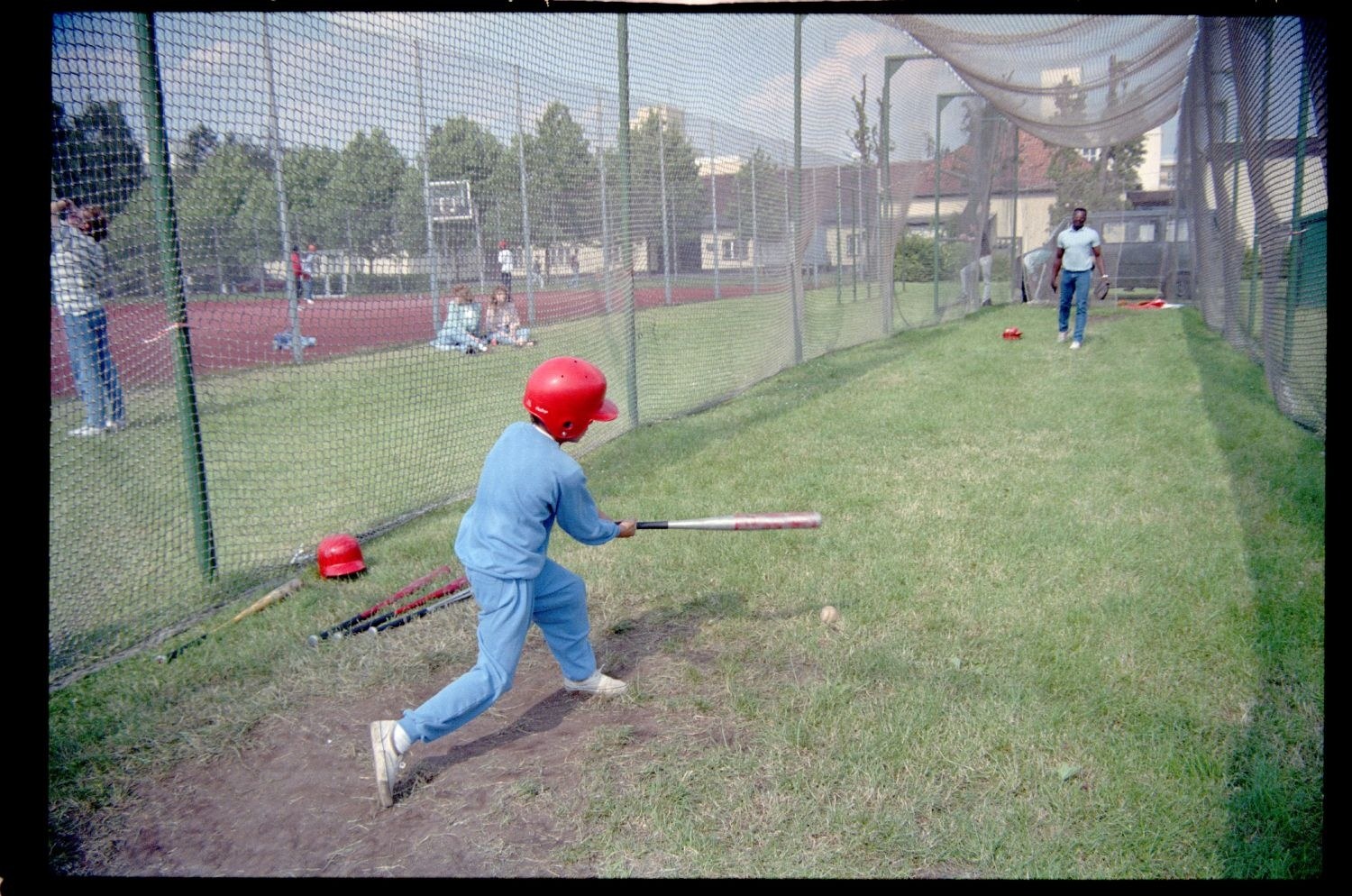 Ein junger Junge in einem blauen Kleid und einem roten Helm schwingt einen Baseballschläger auf einem grünen Feld, mit verstreuten Schlägern und einem Hintergrund aus Bäumen, Gebäuden und einem klaren blauen Himmel.