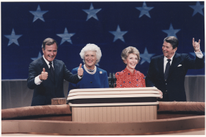 Vier Personen auf einer Bühne, die lächeln und den Daumen hochhalten, mit Mikrofonen auf einem Tisch davor und einer sternförmigen Wand im Hintergrund.