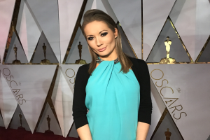 Eine Frau in einem blauen und schwarzen Kleid steht auf einem roten Teppich vor einer Tafel mit der Aufschrift "Oscars 2018 Red Carpet Arrivals" und verschiedenen Skulpturen im Hintergrund.