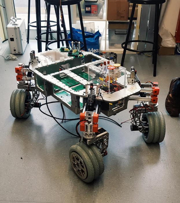 Ein Roboter sitzt auf einem Tisch in einem Raum mit Hockern, Kartons und anderen Gegenständen, mit einem menschlichen Bein auf der rechten Seite.