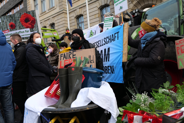Eine Gruppe von Menschen in Masken mit Protestschildern vor einem Lastwagen, mit einem Tisch mit Gegenständen, Topfpflanzen und einem Flaggengebäude im Hintergrund.
