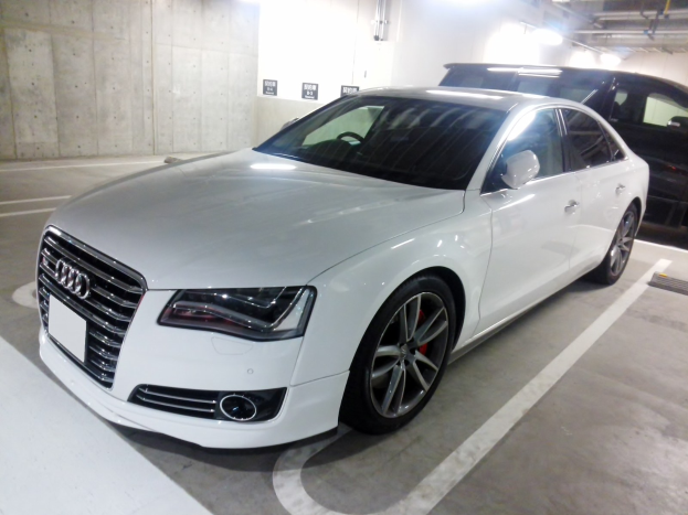 Weißer Audi S8 in einer Parkgarage neben einem schwarzen Auto geparkt, mit Garagenwänden, Deckenrohren und Leuchten im Hintergrund.