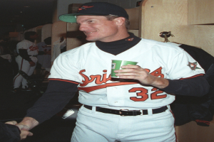 Ein Mann in einem Baseball-Outfit und einer Mütze hält einen Kaffeebecher in einer Umkleidekabine, mit anderen Menschen und einem hölzernen Schrank mit aufgehängten Kleidern im Hintergrund.