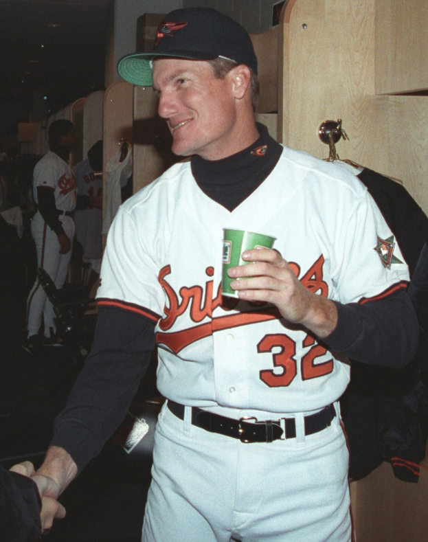 Ein Mann in einem Baseball-Outfit und einer Mütze hält einen Kaffeebecher in einer Umkleidekabine, mit anderen Menschen und einem hölzernen Schrank mit aufgehängten Kleidern im Hintergrund.