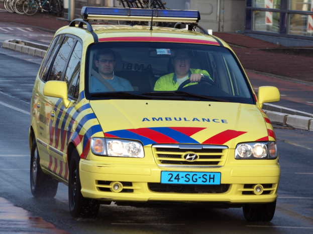 Ein gelber Krankenwagen mit zwei sichtbaren Insassen fährt an parkenden Fahrrädern auf einem Fußweg vorbei, mit einem Glasfenster-fassadengebäude im Hintergrund.