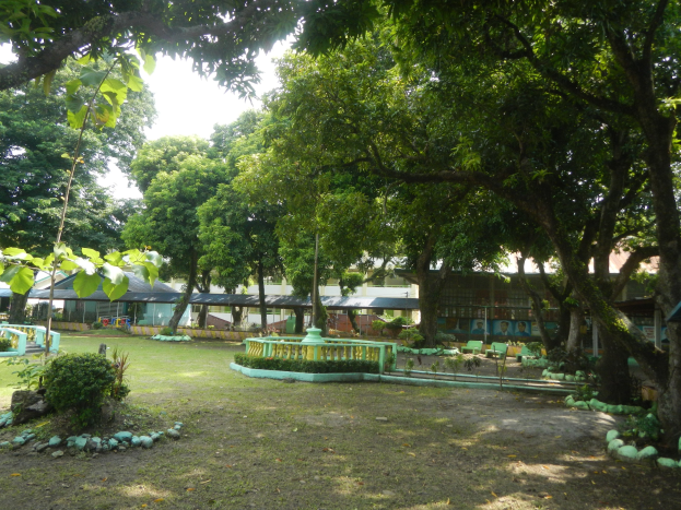 Ein Park mit grünem Gras, Pflanzen, Bäumen, einem zentralen Brunnen, Gebäuden im Hintergrund und einem klaren blauen Himmel.