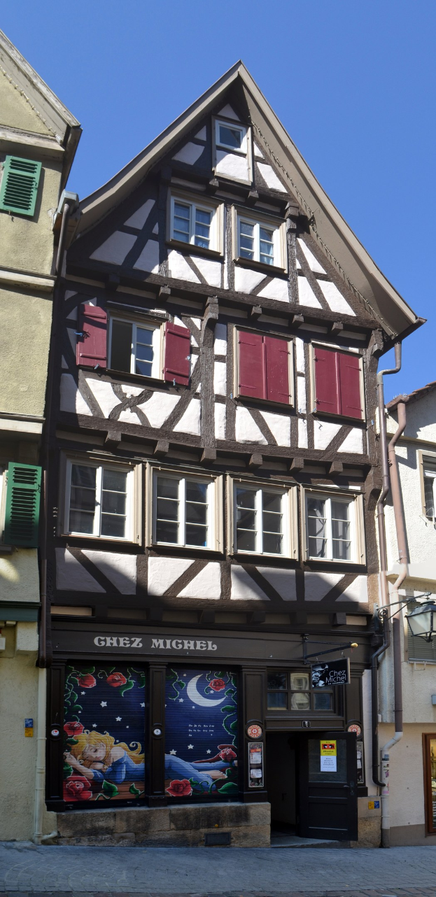 Ein klassisches altes Gebäude mit roten Fensterläden, mehreren Fenstern und einem zeitlosen Design, das sich auf einer Straße unter einem klaren blauen Himmel befindet.