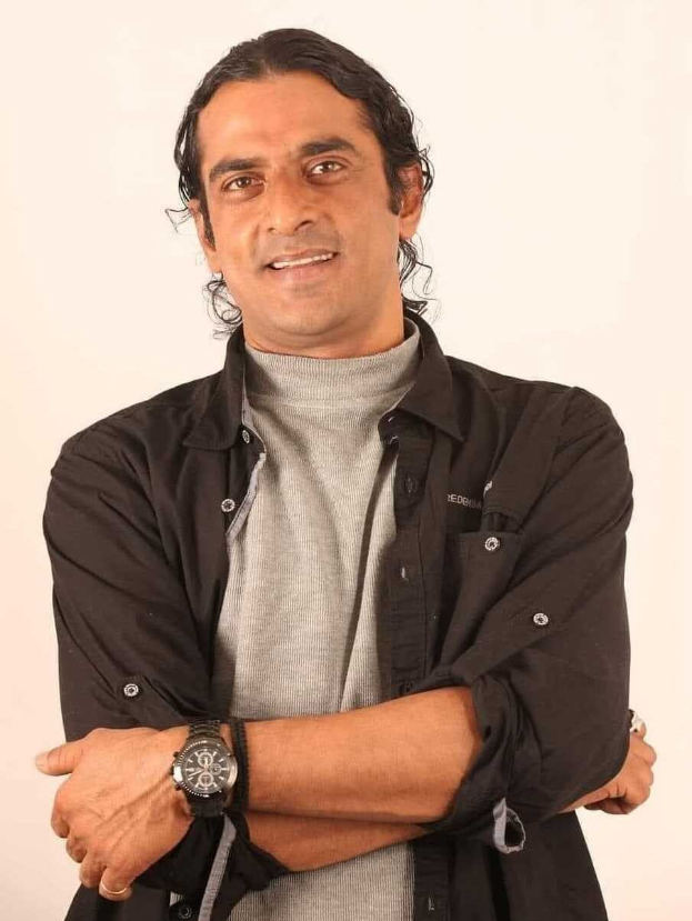 Ein Mann in einem schwarzen und aschfarbenen Kleid mit überkreuzten Armen, der eine Uhr trägt und gegen einen weißen Hintergrund steht, der nachdenklich erscheint.