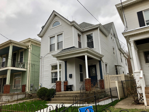 Reihe von Häusern mit Fenstern, Türen, Säulen, Geländern, Stufen und Dächern, umgeben von einem Metallzaun, Gras, Pflanzen, Bäumen und einem bewölkten Himmel, mit einem Vermietungsschild, das die Adresse und die Listen Details anzeigt.