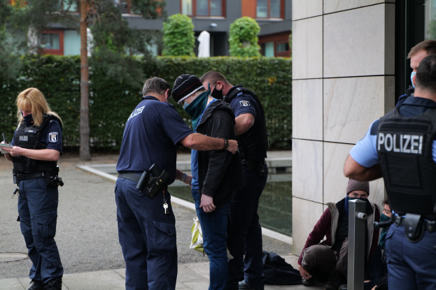 Eine Gruppe von Polizeibeamten steht vor einem Gebäude, während eine Frau ein Buch und einen Stift hält, umgeben von sitzenden und stehenden Personen und Bäumen sowie Gebäuden im Hintergrund.