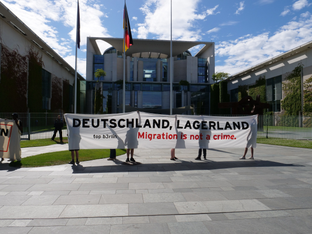 Eine Gruppe von Menschen mit einem Transparent mit der Aufschrift "Deutschland, Lagerland Migration ist kein Verbrechen" vor einem umzünten Gebäude mit Bäumen und Fahnen.