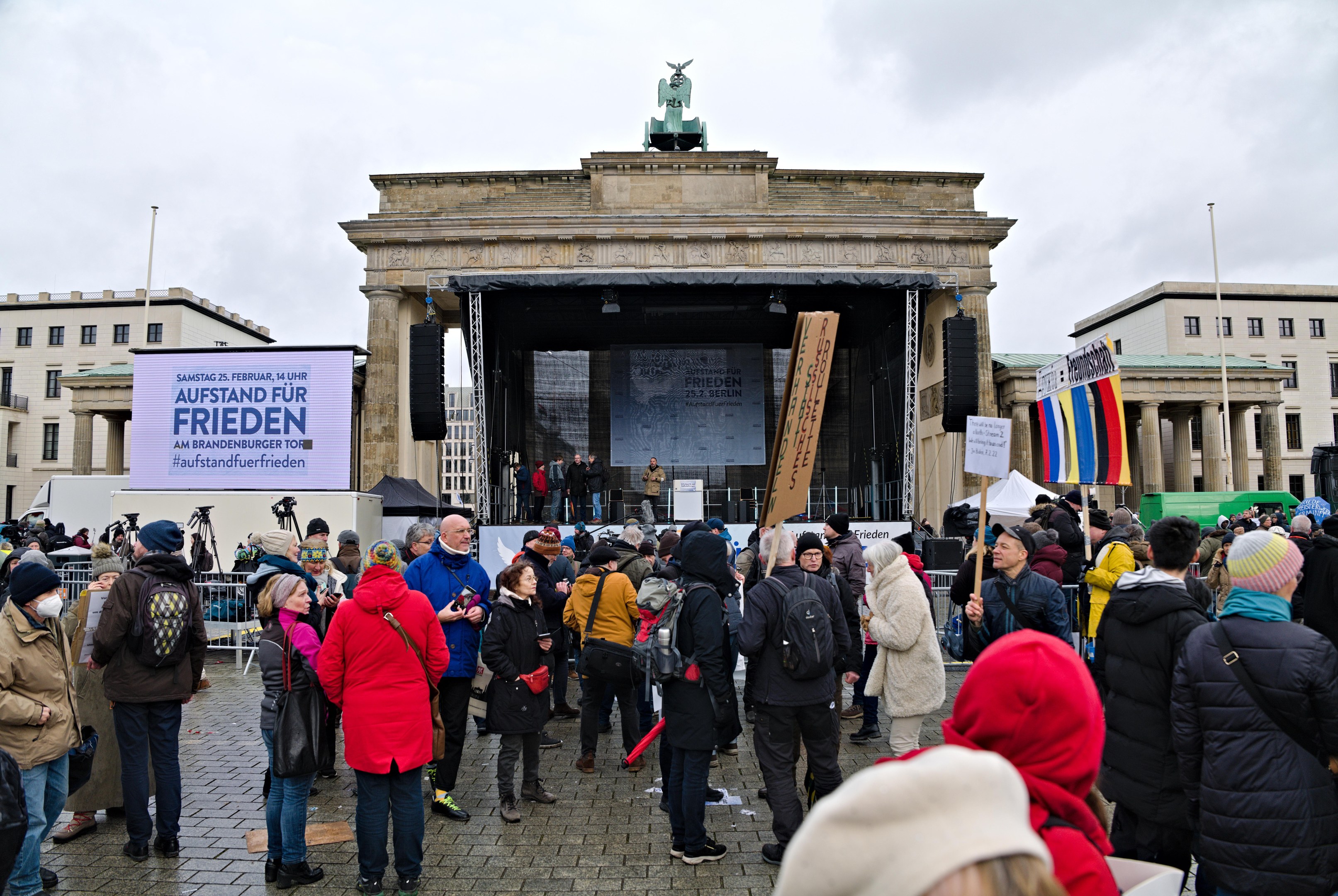 Menge von Menschen versammelt vor einem Gebäude mit einer Bühne, auf der Redner und ein Bildschirm zu sehen sind, umgeben von Fahnen und Transparenten mit Text, in Berlin während einer Demonstration.