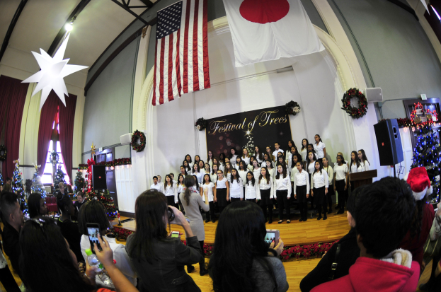 Eine Gruppe von Menschen auf einer Bühne vor einem Publikum mit Weihnachtsdekoration, einem Podium, Lautsprechern, einer Texttafel, Vorhängen, Flaggen und Deckenleuchten.