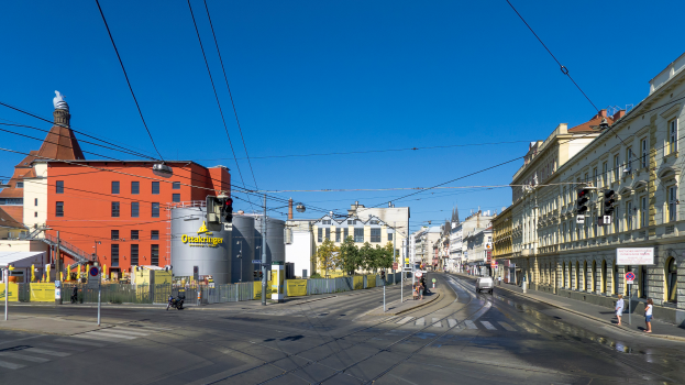 Eine Straßenszene in der Stadt mit Gebäuden, Straßenlaternen, Verkehrszeichen, Fahrzeugen, Fußgängern, Bäumen und einem Himmel im Hintergrund.