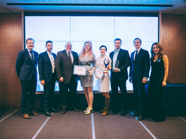 Gruppe von strahlenden Menschen mit Urkunden vor einem Bildschirm, neben einem schwarz bedeckten Tisch mit einem Hintergrundobjekt an der Wand, unter Deckenlampen, Gewinner der Internationalen Wirtschaftsauszeichnungen 2018.