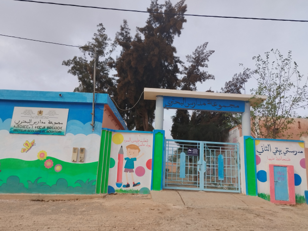 Blau-weißes Gebäude mit Wandgemälde, Eingangstor, Namensschild und umliegenden Häusern, Bäumen, Strommast und bewölktem Himmel; das Gebäude ist der Eingang zu einer Kinder Schule.