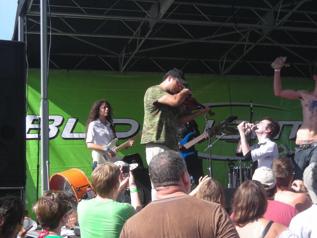 Eine Gruppe von Musikern, die auf der Bühne mit verschiedenen Instrumenten spielen, ein großer schwarzer Lautsprecher links und ein Publikum vor einem Podium mit einer grünen Fahne im Hintergrund.