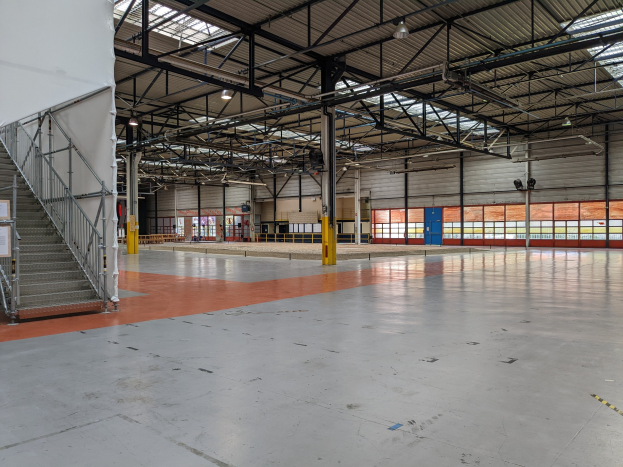 Großes Industriegebäude mit zahlreichen Fenstern, Metallstützen, einer Treppe mit Geländer, einer Tafel, Deckenleuchten und zusätzlichen Stützen.