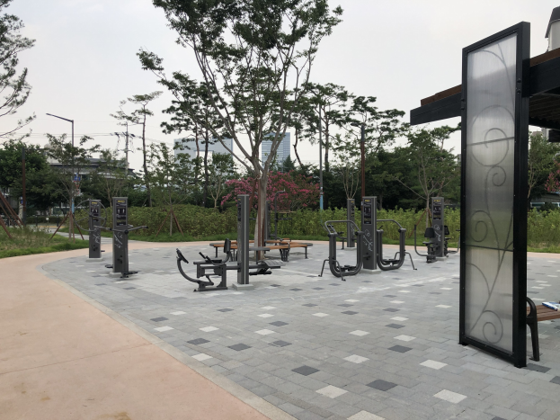 Outdoor park featuring fitness equipment, benches, trees, plants, grass, poles, lights, wires, and buildings under a sky backdrop.
