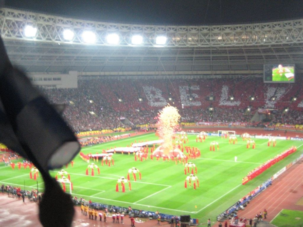 Ein Stadion mit Flutlicht, Eisengittern, Zuschauern, stehenden Personen, Werbetafeln, Bildschirmen, einer Überwachungskamera, Fahnen und Dekorationselementen.