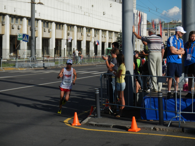 Mann rennt öffentlich durch eine Menschenmenge, mit Verkehrskegeln, Barrieren, Pfosten, Fahnen, Schilder, Geländern, Gebäuden, Bäumen und einem bewölktem Himmel im Hintergrund.