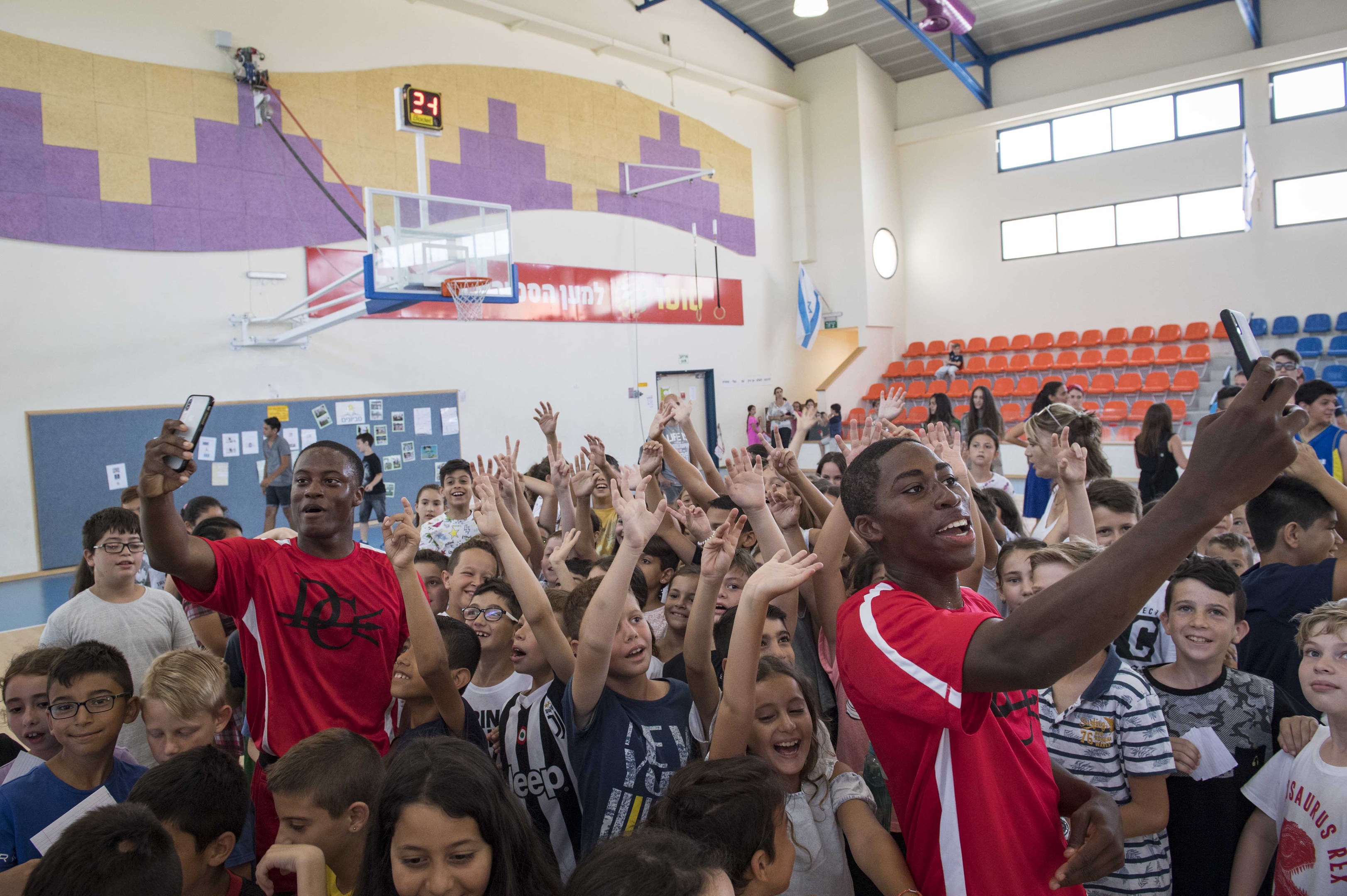 Kinder vor einem Basketballfeld an einem Camp stehend, einige halten Telefone; eine Wandtafel mit Papieren, eine Uhr, ein Torpfosten, ein Basketballkorb, Deckenlichter, Stühle und Fenster sind zu sehen.