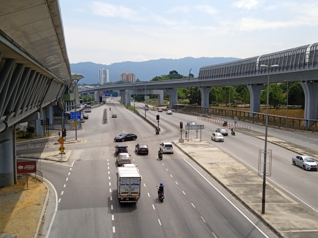 Eine vielbefahrene Autobahn mit mehreren Fahrspuren neben einer Brücke, mit Straßenlaternen, Ampeln, Verkehrszeichen, Bäumen, Gebäuden, Hügeln und einem bewölkten Himmel.