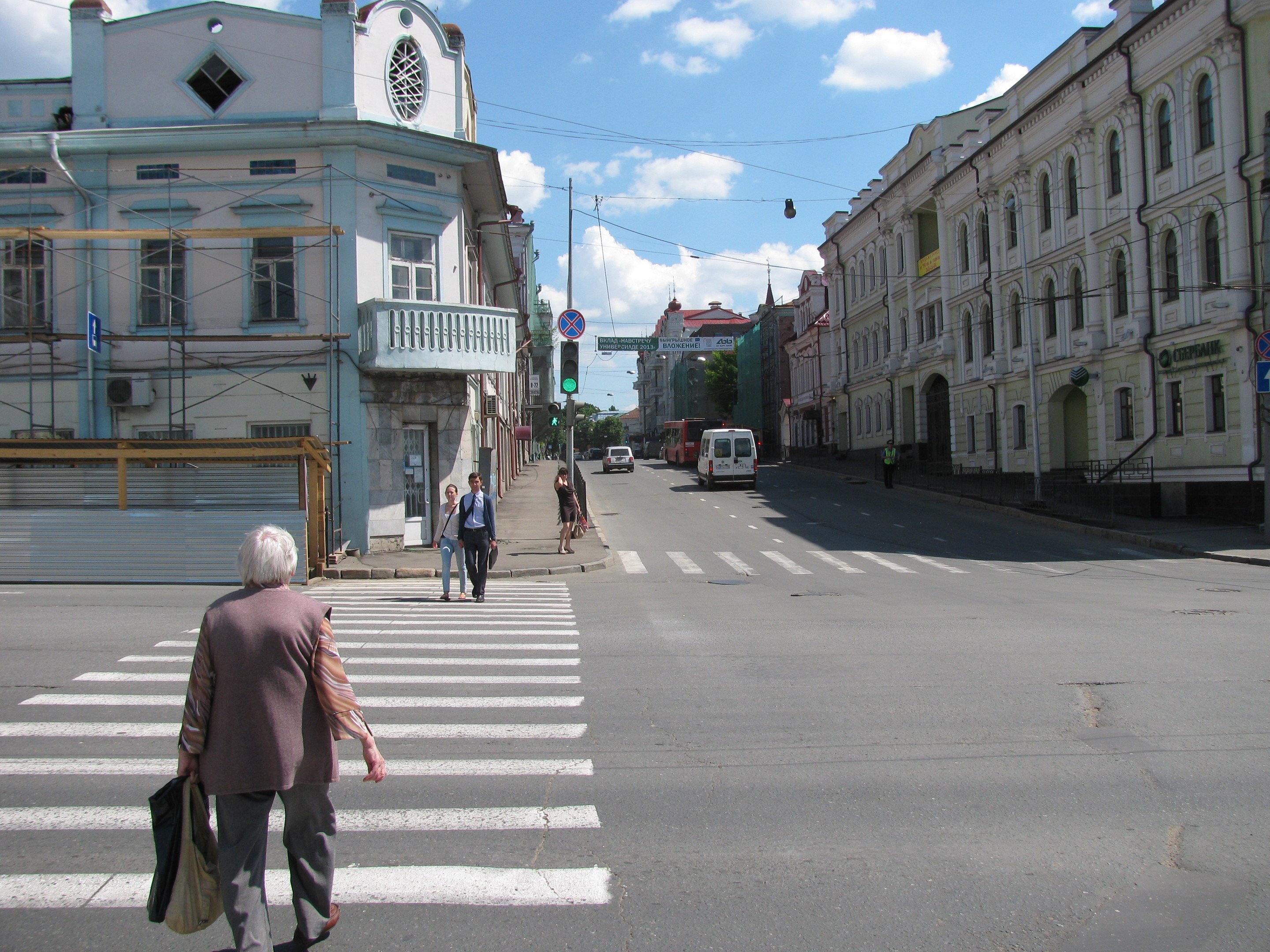 Alte Frau überquert eine Straße vor einem Gebäude, mit Fußgängern, Fahrzeugen, Schildern, Strommasten, Bäumen und einem bewölkten Himmel im Hintergrund.